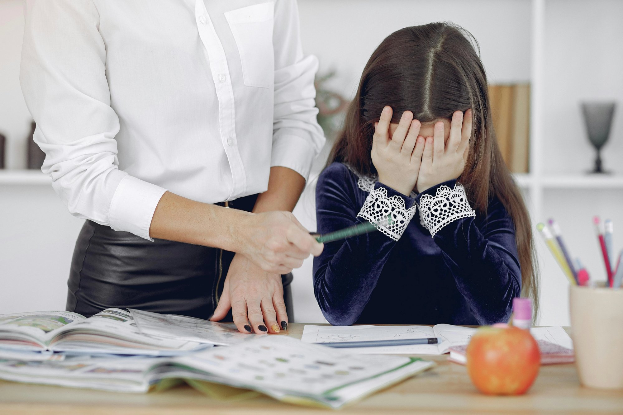Child covering her face with her hands over schoolwork while an adult stands nearby — representing the frustration of an unidentified or undersupported learning disability